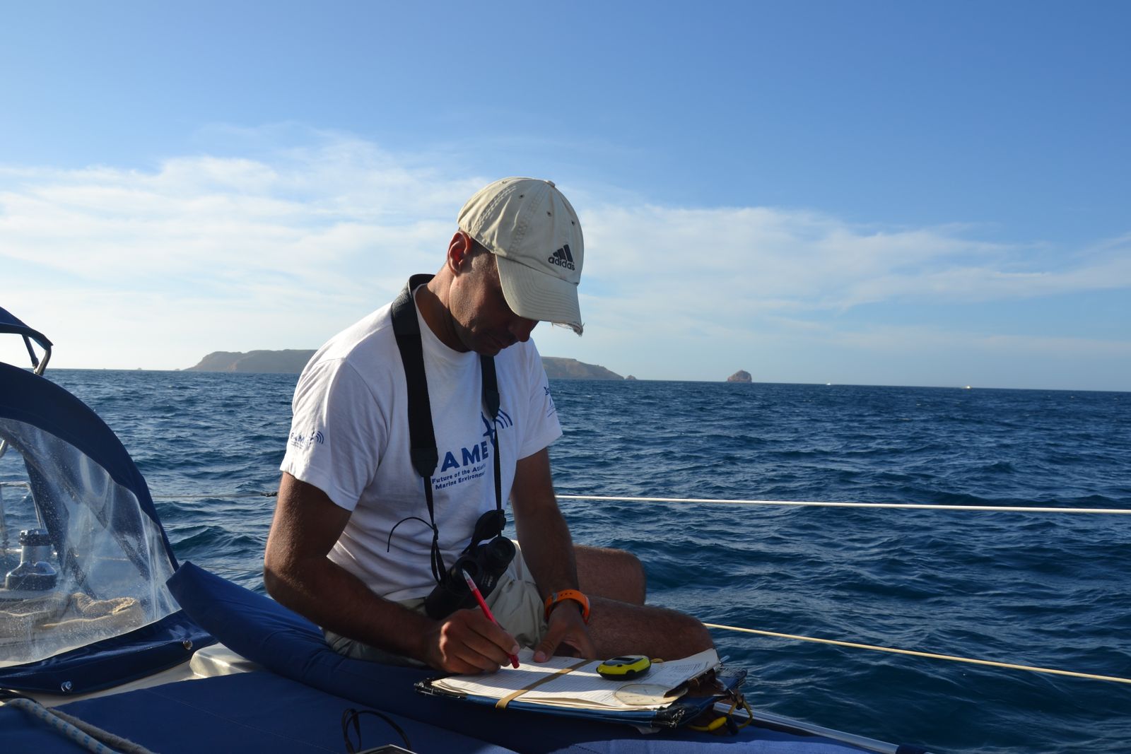 Investigador a bordo, com binóculos ao pescoço, a registar observações no âmbito do censo ESAS. Ao fundo, observa-se a ilha da Berlenga e, no barco, parte do equipamento utilizado no censo, incluindo um cronómetro e um GPS.