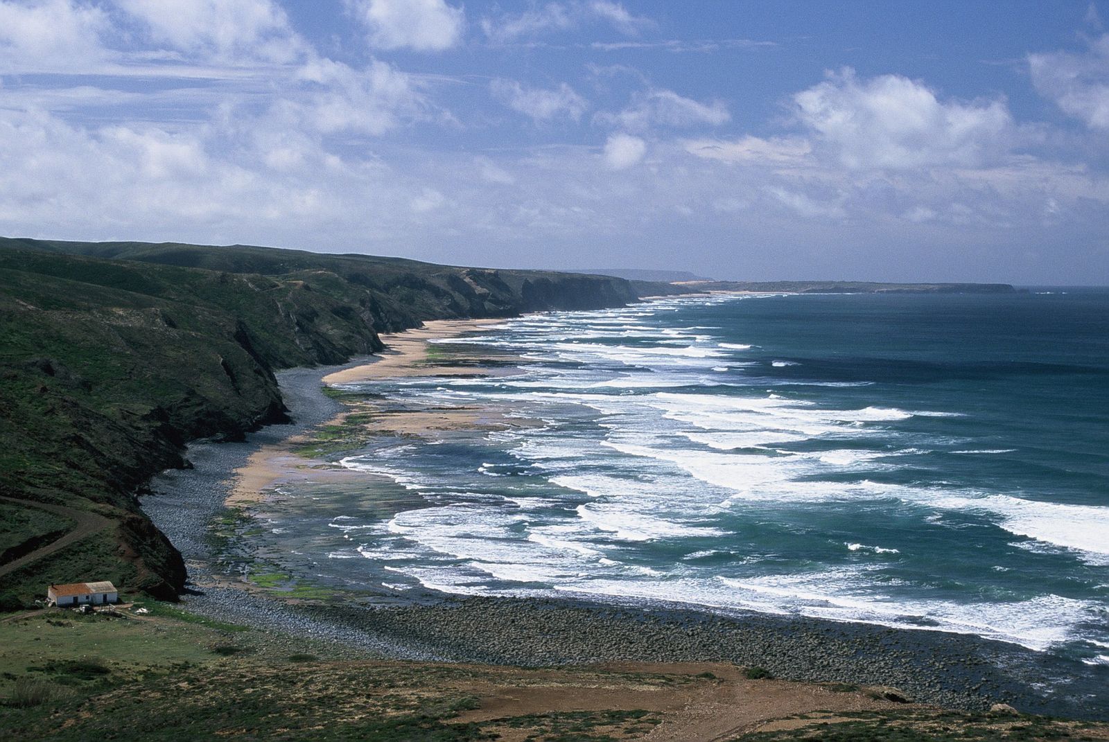 Linha de costa com falésias acentuadas, uma zona de areal e substrato rochoso. Nota-se a espuma branca de um mar bastante encrespado.